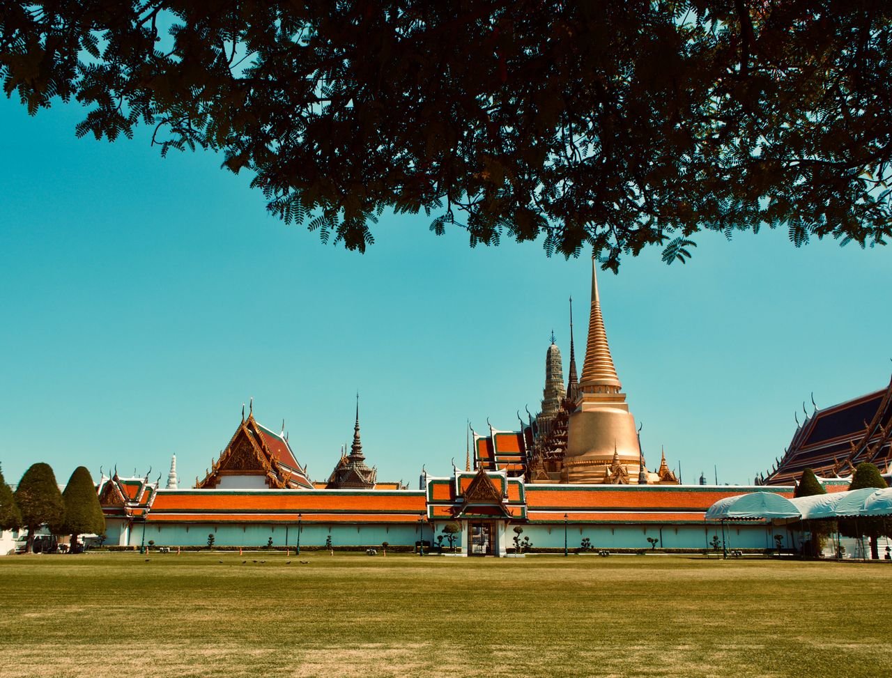 Wide-angle view of the Grand Palace in Bangkok with blue sky and lush trees.