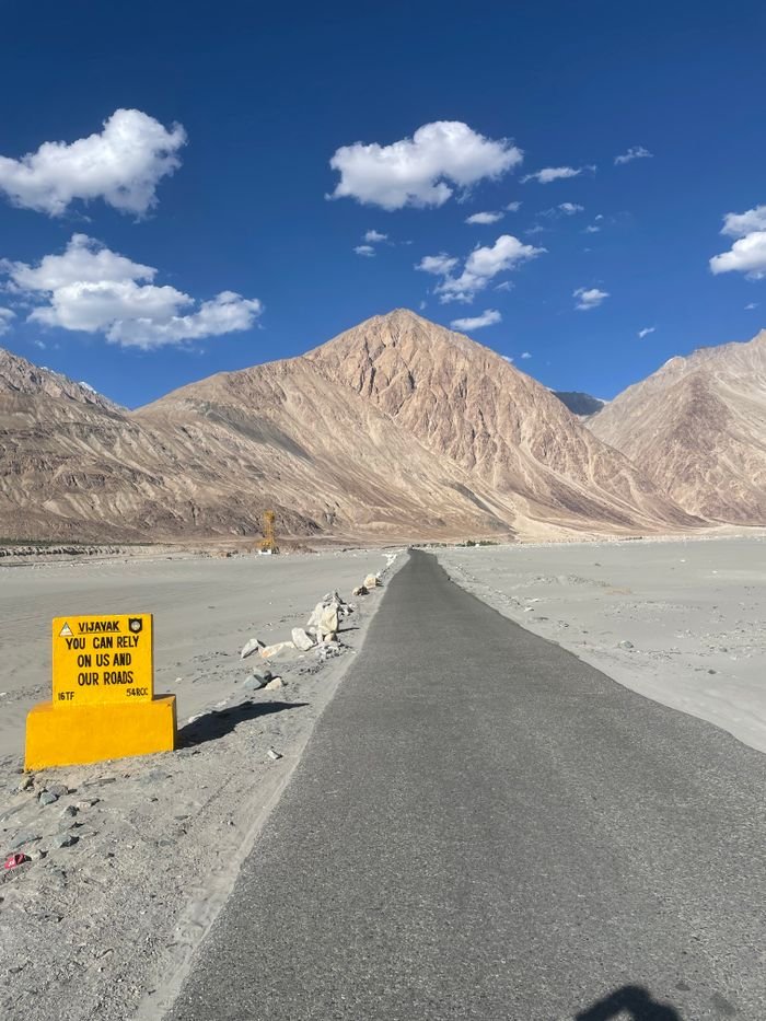 A paved road leads to majestic mountains in Ladakh, India, under a clear blue sky with fluffy clouds.